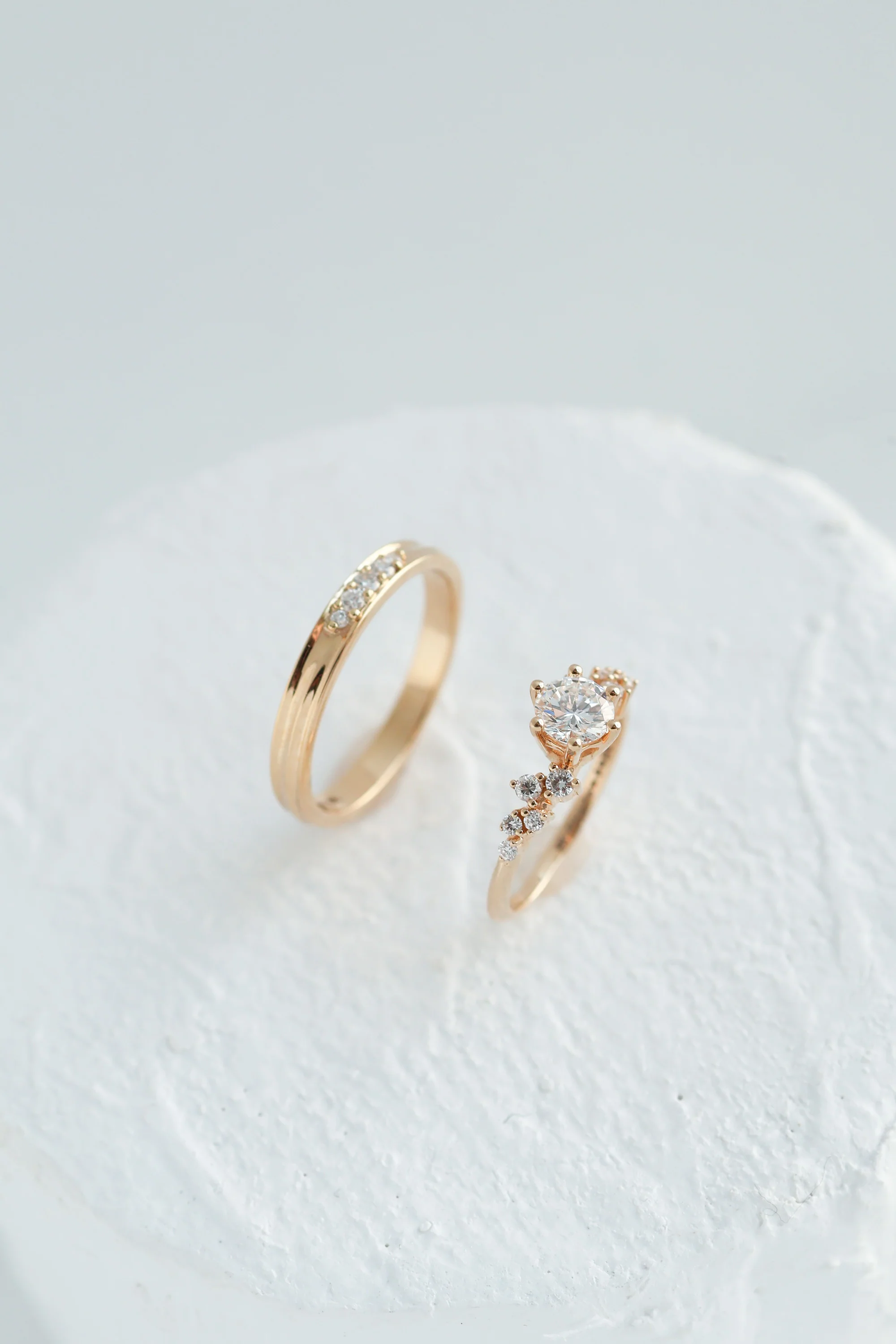 Two gold rings, one a plain band set with pavé diamonds, the other a solitaire with a side cluster, on a white textured surface